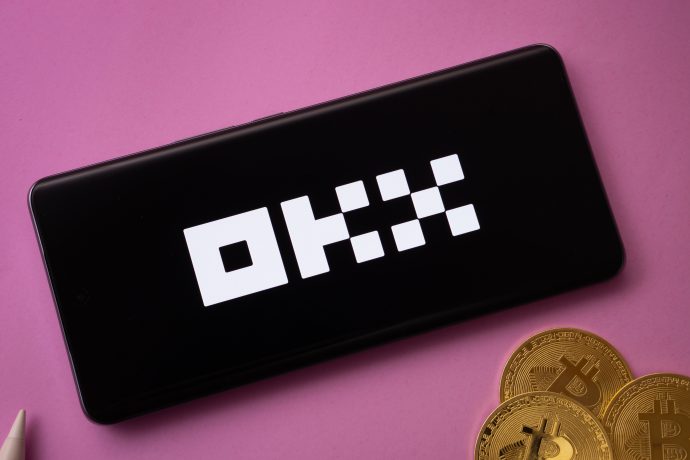 Ein Smartphone mit dem OKX-Logo auf dem Bildschirm liegt neben mehreren physischen Bitcoin-Münzen auf einer rosa Oberfläche und unterstreicht die Verbindung von OKX zur Welt der Kryptowährungen.