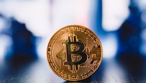 Ein goldfarbener physischer Bitcoin steht aufrecht auf einer reflektierenden Oberfläche mit unscharfem Hintergrund.