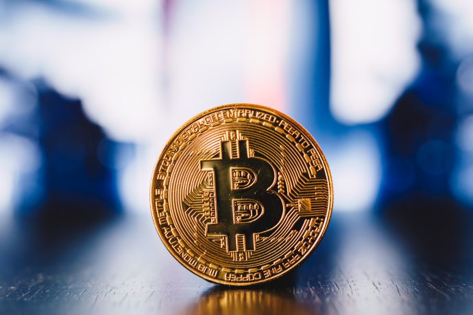 Ein goldfarbener physischer Bitcoin steht aufrecht auf einer reflektierenden Oberfläche mit unscharfem Hintergrund.