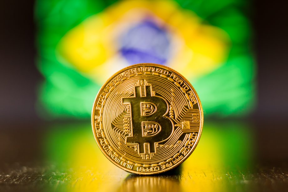 Eine goldene Bitcoin-Münze steht aufrecht auf einer reflektierenden Oberfläche mit einer verschwommenen brasilianischen Flagge im Hintergrund.
