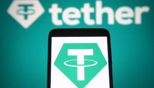 Auf einem Smartphone-Bildschirm wird das Tether-Logo angezeigt, wobei im Hintergrund ein größeres Tether-Logo und das Wort „Tether“ sichtbar sind, was das wachsende Interesse inmitten der Diskussionen über einen möglichen Börsengang unterstreicht.