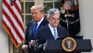 Donald Trump steht hinter Jerome Powell, der an einem Podium mit dem Siegel des Präsidenten spricht; im Hintergrund sind die US-Flagge und die Flagge des Präsidenten zu sehen.