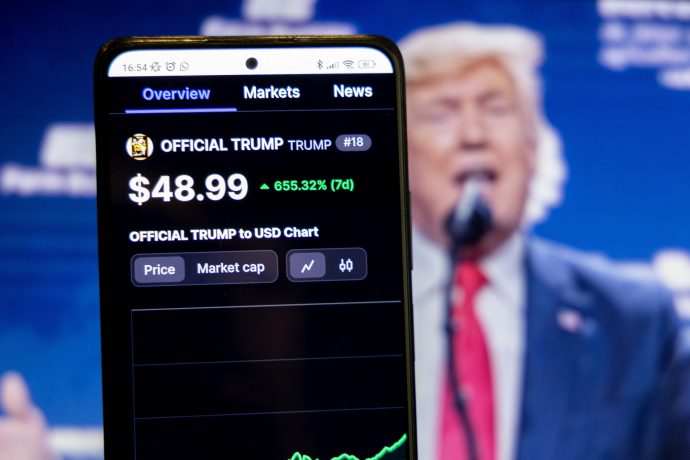 Ein Smartphone zeigt den Preis einer Kryptowährung namens OFFICIAL TRUMP mit 48,99 $ an, der wöchentlich um 655,32 % gestiegen ist; im unscharfen Hintergrund spricht ein Mann.