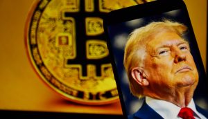 Ein Smartphone mit einem Foto von Donald Trump wird vor ein großes Krypto-Bitcoin-Symbol auf einem Bildschirm im Hintergrund gehalten.