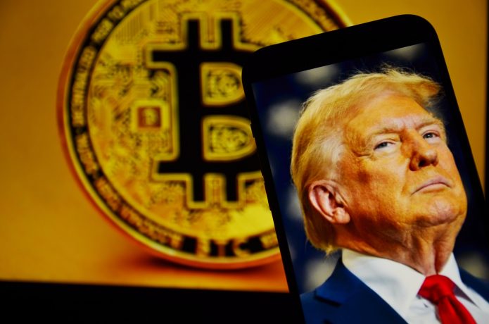 Ein Smartphone mit einem Foto von Donald Trump wird vor ein großes Krypto-Bitcoin-Symbol auf einem Bildschirm im Hintergrund gehalten.