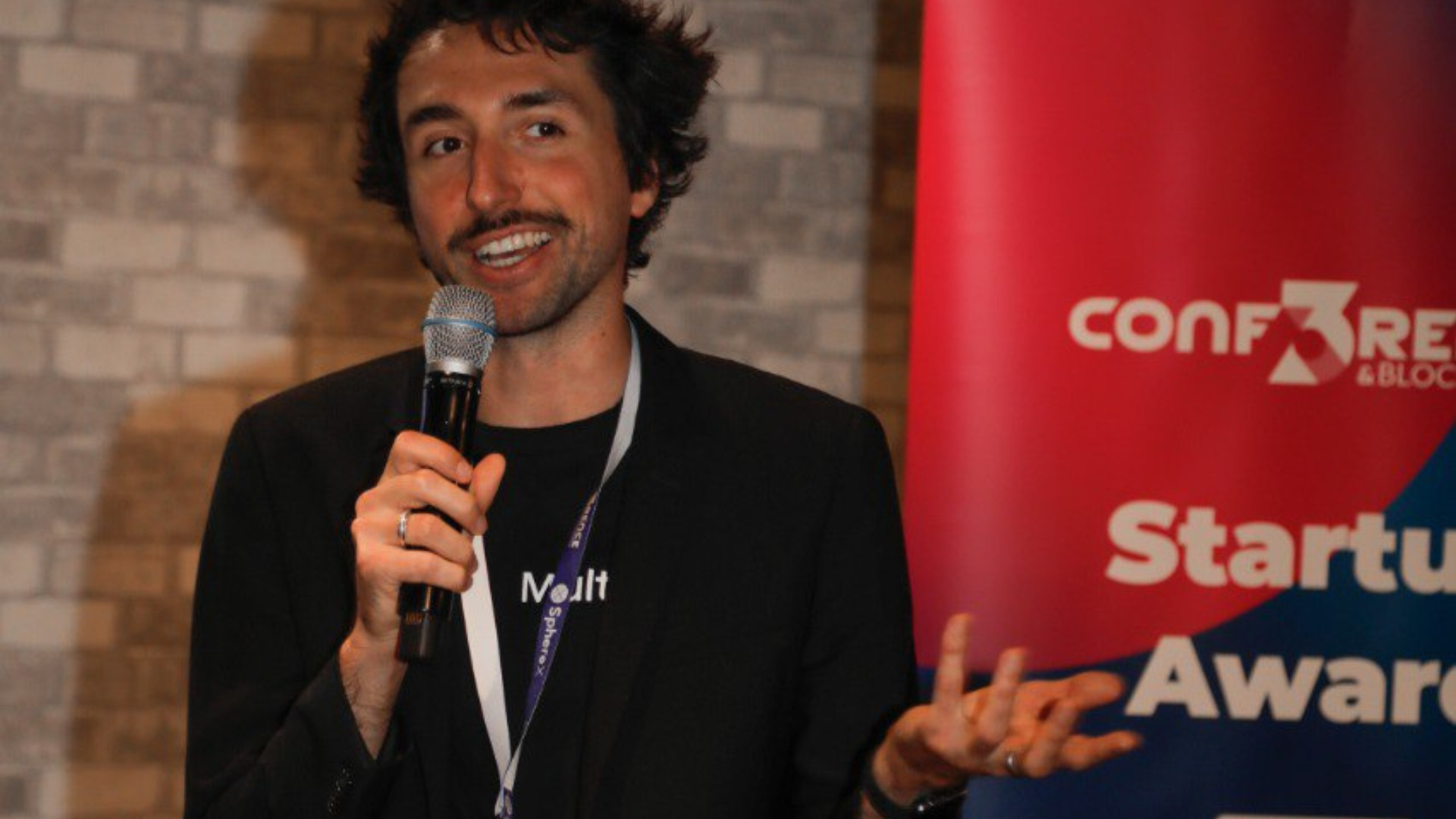 1 Ein Mann hält ein Mikrofon in der Hand und spricht bei der Startup Award-Veranstaltung in Dortmund vor einer Backsteinmauer und einem Banner mit der Aufschrift Startup Awards.
