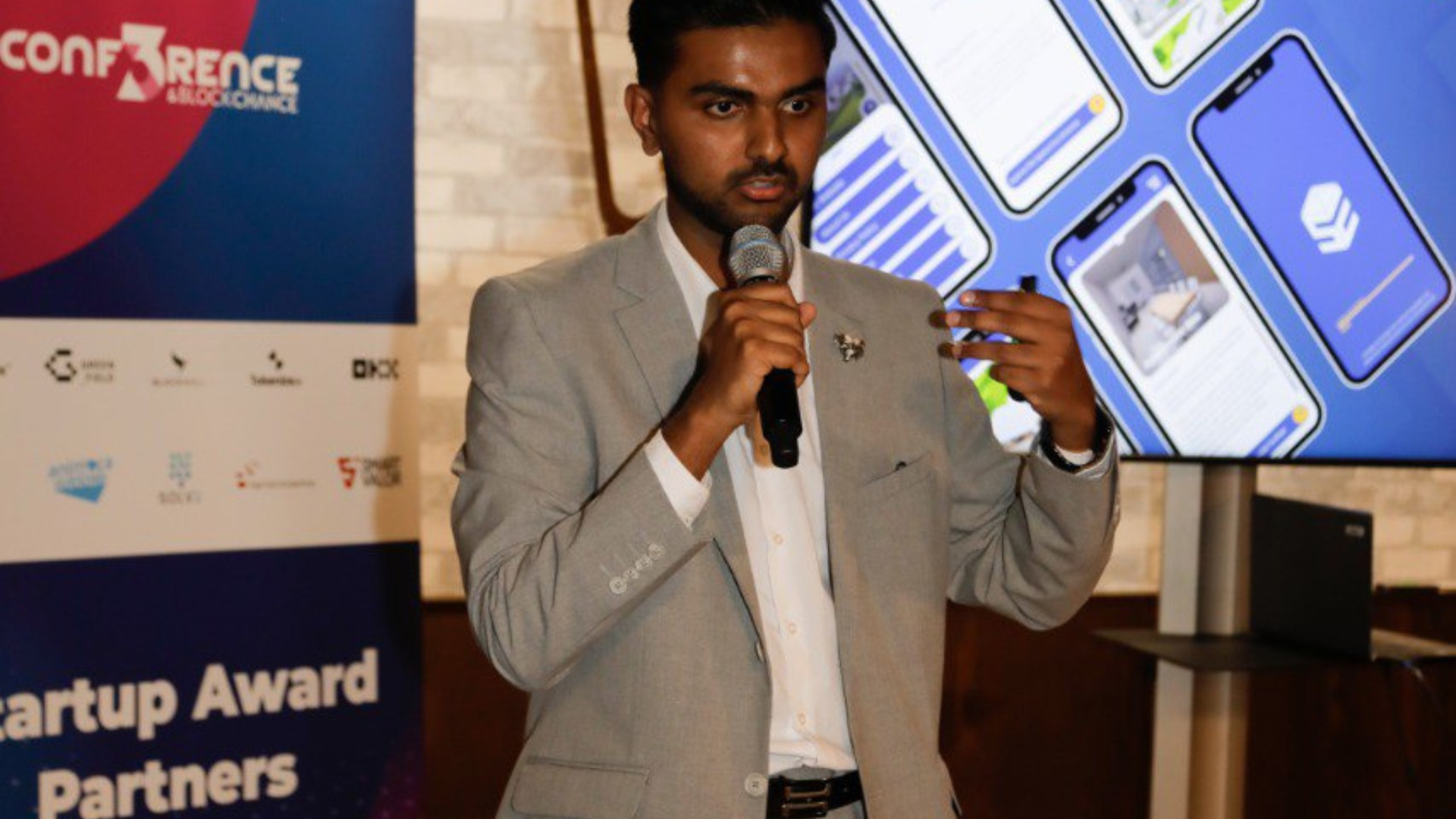 2 Ein Mann in einem grauen Anzug hält ein Mikrofon in der Hand und spricht während einer Präsentation beim Start-up Award in Dortmund. Im Hintergrund sind ein digitaler Bildschirm und ein Veranstaltungsbanner zu sehen.