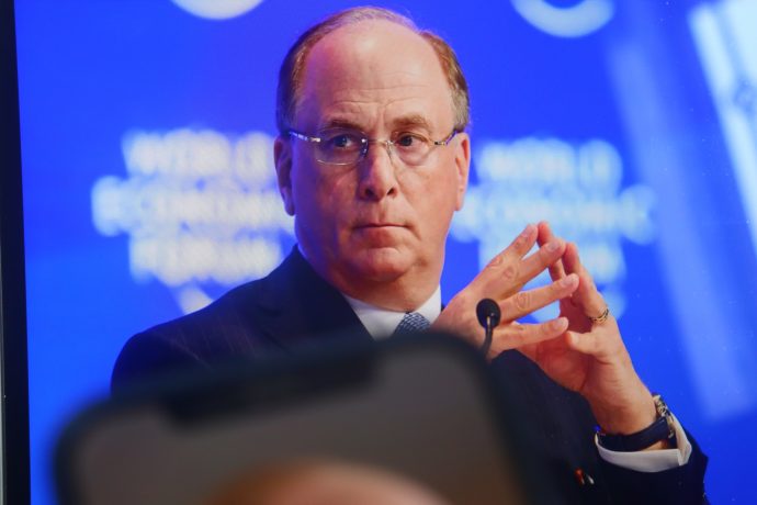Ein Mann im Anzug mit Brille, möglicherweise ein leitender Angestellter von BlackRock, sitzt vor einem blauen Hintergrund und schaut ernst mit verschränkten Fingern.