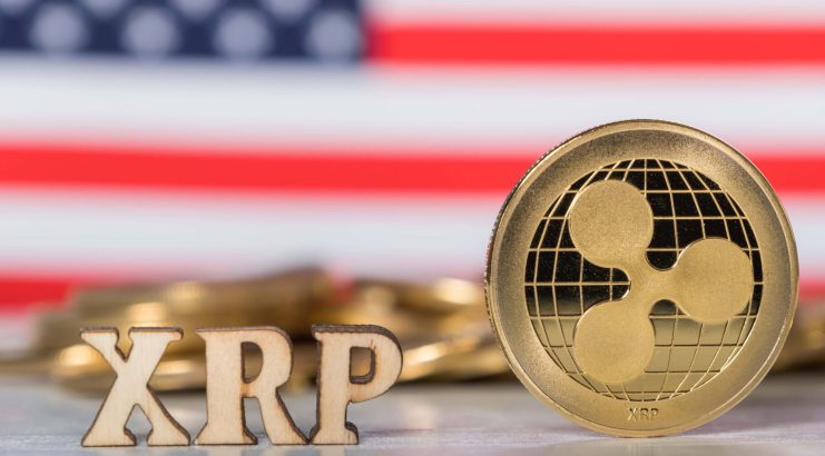 Eine goldene XRP-Münze steht aufrecht neben hölzernen XRP-Buchstaben, mit einer verschwommenen amerikanischen Flagge im Hintergrund, die die Bedeutung von XRP hervorhebt.