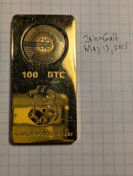 Barren Ein Casascius Bitcoin Barren, vergoldet und mit einem Hologramm auf der Oberseite als 100 BTC gekennzeichnet, liegt auf Millimeterpapier neben einem handgeschriebenen Text: John Galt 13. Mai 2025.