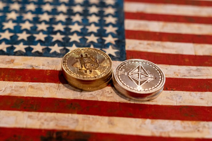 Zwei physische Kryptowährungsmünzen, darunter ein Bitcoin, werden auf eine Flagge der Vereinigten Staaten gelegt.