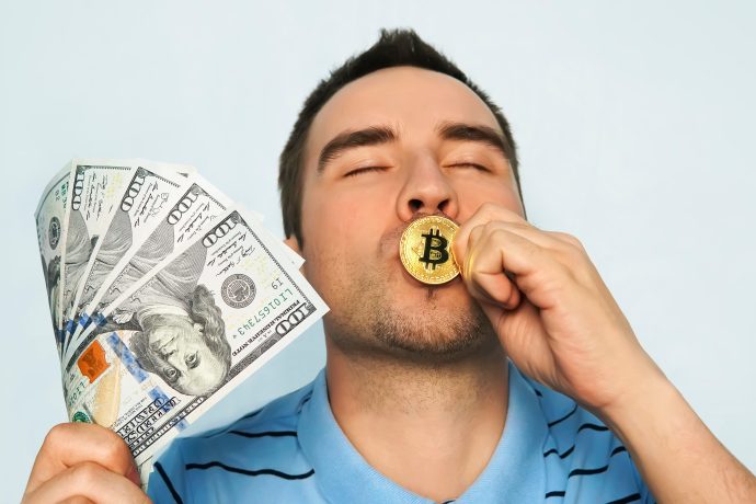 Ein Mann hält mehrere US-Hundert-Dollar-Scheine in der einen Hand und küsst mit der anderen eine goldene Bitcoin-Münze, wobei er vor einem schlichten hellen Hintergrund stolz seine Liebe zu Bitcoin zur Schau stellt.