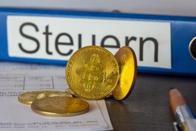 Mehrere physische Bitcoin-Token liegen auf Dokumenten neben einem blauen Ordner mit der Aufschrift "Steuern" und einem Bleistift, der die Überschneidung von Krypto-Vermögenswerten und Steuervorschriften verdeutlicht.