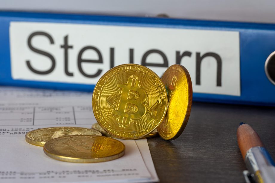Mehrere physische Bitcoin-Token liegen auf Dokumenten neben einem blauen Ordner mit der Aufschrift "Steuern" und einem Bleistift, der die Überschneidung von Krypto-Vermögenswerten und Steuervorschriften verdeutlicht.