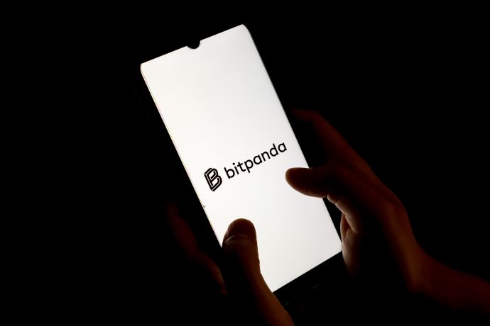 Eine Person hält ein Smartphone in der Hand, auf dem das Bitpanda-Logo auf einem weißen Bildschirm vor einem dunklen Hintergrund angezeigt wird, was die schlanke und moderne Benutzeroberfläche von Bitpanda hervorhebt.
