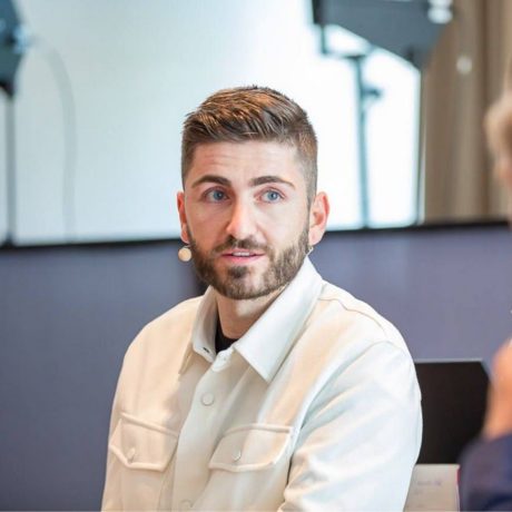 Ein Mann mit kurzen braunen Haaren und einem Bart, der ein weißes Hemd und ein Headset-Mikrofon trägt, spricht in einem Innenraum, in dem unscharfe Hintergrundgeräte zu sehen sind - möglicherweise spricht er zum Publikum, nachdem er einen 2025 Influencer Award gewonnen hat.