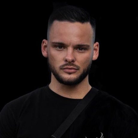 Ein Mann mit kurzen dunklen Haaren, gestutztem Bart und ernstem Gesichtsausdruck trägt ein schwarzes Hemd und eine Umhängetasche vor einem schlichten schwarzen Hintergrund und zeigt damit das selbstbewusste Auftreten eines für den Influencer Award 2025 nominierten Kandidaten.