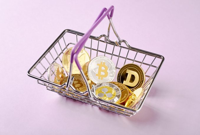 Ein kleiner Einkaufskorb mit verschiedenen Kryptowährungsmünzen, darunter Bitcoin, Ethereum, Ripple und Dogecoin tokenisierte Indizes, auf einem hellrosa Hintergrund.