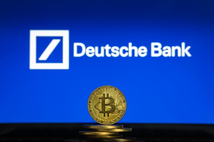 Eine Bitcoin-Münze steht aufrecht vor einem Stapel Münzen, während das Logo der Deutschen Bank auf einem blauen Bildschirm im Hintergrund angezeigt wird.