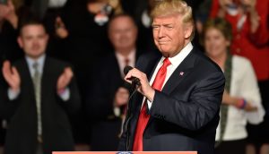 Donald Trump steht im Anzug und mit roter Krawatte auf einem Podium mit einem Mikrofon und spricht zu der Menge über Bitcoin, während die Leute im Hintergrund applaudieren.