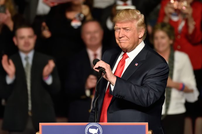 Donald Trump steht im Anzug und mit roter Krawatte auf einem Podium mit einem Mikrofon und spricht zu der Menge über Bitcoin, während die Leute im Hintergrund applaudieren.