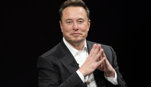 Elon Musk sitzt auf einem Podium, die Hände zusammengefaltet