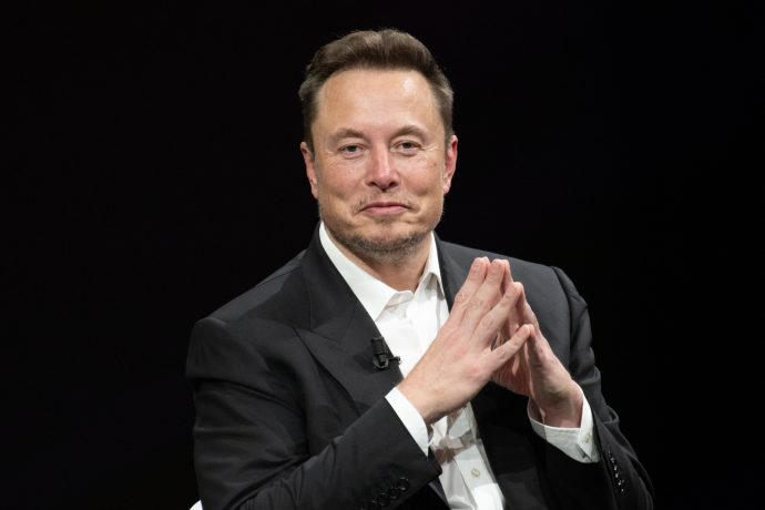 Elon Musk sitzt auf einem Podium, die Hände zusammengefaltet