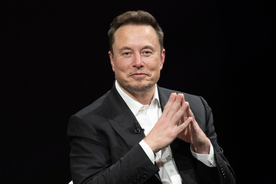 Elon Musk sitzt auf einem Podium, die Hände zusammengefaltet