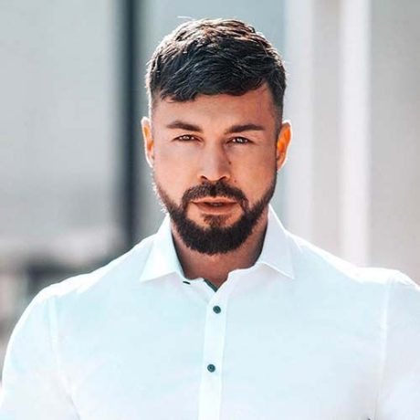 Ein Mann mit kurzen braunen Haaren und einem Bart, der ein weißes Button-up-Hemd trägt, steht vor einem unscharfen Hintergrund im Freien und ist bereit, am Influencer Award 2025 teilzunehmen.