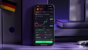 Ein Smartphone zeigt die Kraken-App für den Handel mit Kryptowährungen mit einem Bitcoin-Kursdiagramm an, das auf einem lilafarbenen Schreibtisch neben Büchern und einer deutschen Flagge steht.