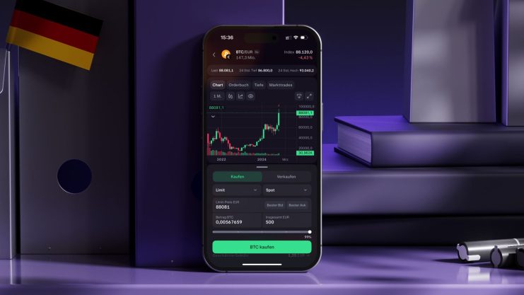Ein Smartphone zeigt die Kraken-App für den Handel mit Kryptowährungen mit einem Bitcoin-Kursdiagramm an, das auf einem lilafarbenen Schreibtisch neben Büchern und einer deutschen Flagge steht.