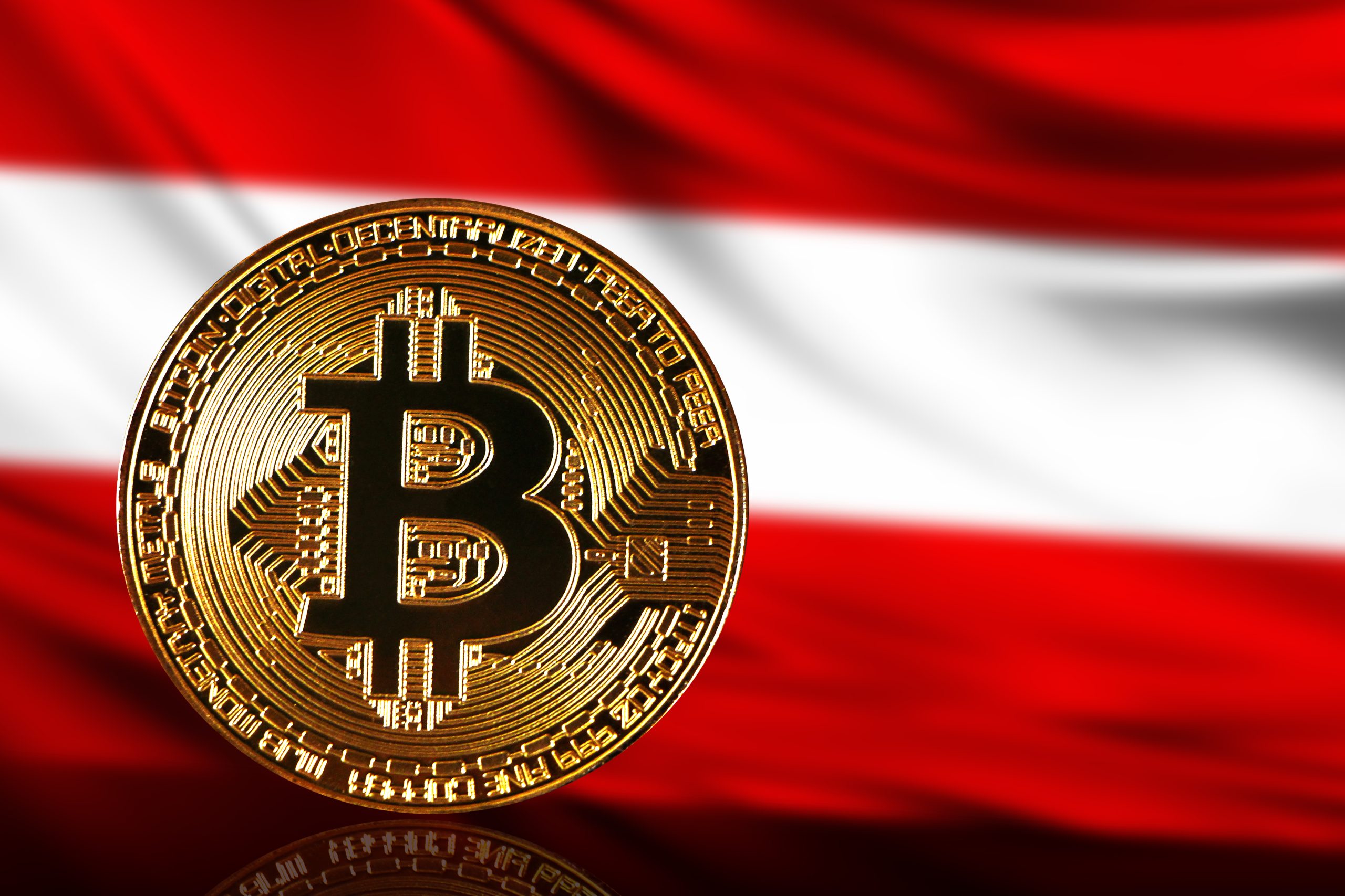 Eine goldene Bitcoin-Münze, die Krypto symbolisiert, ist vor der österreichischen Flagge mit ihren charakteristischen rot-weiß-roten Querstreifen zu sehen.