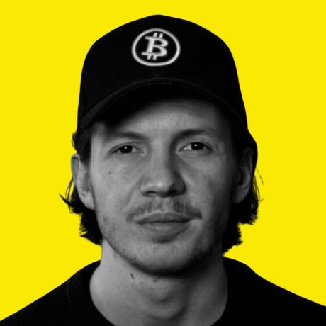 Ein Mann, der eine schwarze Kappe mit einem Bitcoin-Logo trägt, blickt in die Kamera und posiert vor einem leuchtend gelben Hintergrund - perfekt, um für den Influencer Award 2025 zu werben.