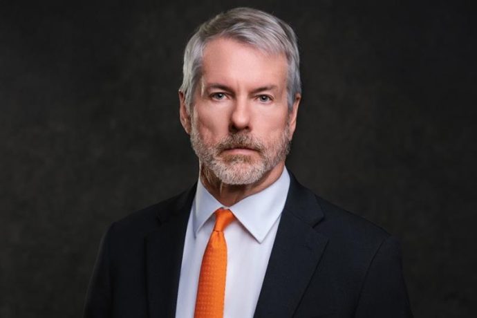 Michael Saylor, der Gründer von MicroStrategy, mit grauem Haar und Bart, der einen schwarzen Anzug, ein weißes Hemd und eine orangefarbene Krawatte trägt, posiert vor einem dunklen Hintergrund.