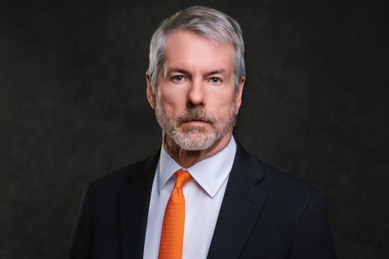 Michael Saylor, der Gründer von MicroStrategy, mit grauem Haar und Bart, der einen schwarzen Anzug, ein weißes Hemd und eine orangefarbene Krawatte trägt, posiert vor einem dunklen Hintergrund.