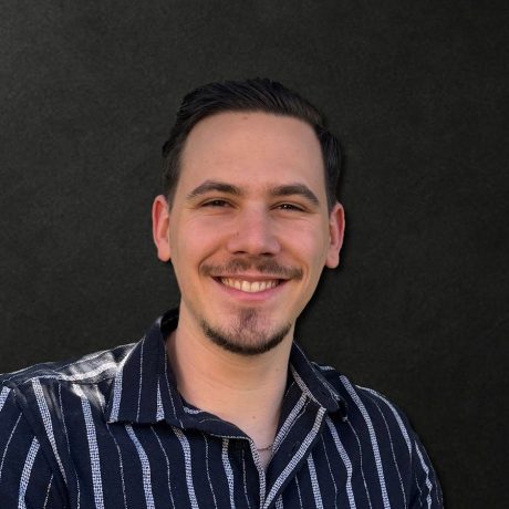 Ein Mann mit kurzen dunklen Haaren und gestutztem Bart, der ein schwarz-weiß gestreiftes Hemd trägt, lächelt selbstbewusst vor einem schlichten dunklen Hintergrund und verkörpert die Ausstrahlung eines Influencer Award 2025-Gewinners.