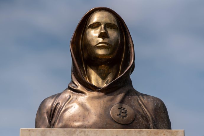 Eine Bronzestatue einer vermummten Figur, die Satoshi Nakamoto symbolisiert, mit einem Bitcoin-Emblem auf der Brust, vor einem wolkenverhangenen Hintergrund.
