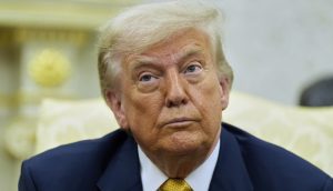 Trump in einem Anzug mit gelber Krawatte, der an Fed-Chef Jerome Powell erinnert, sitzt in einem Raum und blickt mit neutraler Miene leicht nach oben.