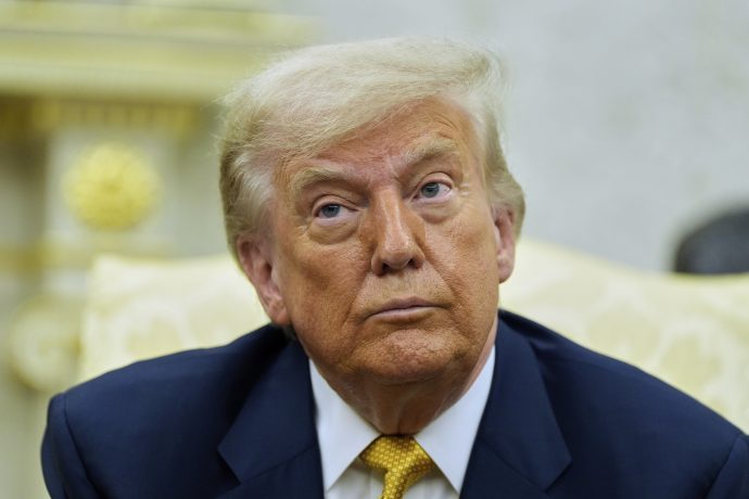 Trump in einem Anzug mit gelber Krawatte, der an Fed-Chef Jerome Powell erinnert, sitzt in einem Raum und blickt mit neutraler Miene leicht nach oben.