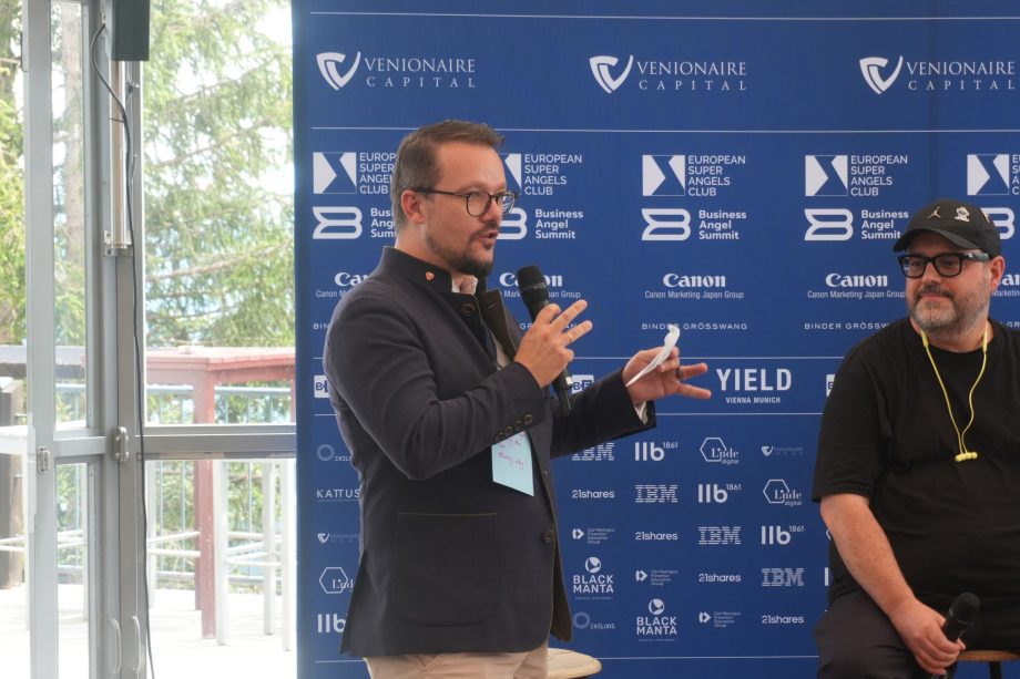 berthold keynote Ein Mann, der in ein Mikrofon spricht und Notizen in der Hand hält, steht neben einem anderen sitzenden Mann vor einem blauen Veranstaltungshintergrund mit verschiedenen Sponsorenlogos und spricht über Konzepte wie tokenisierte Indizes und ETF.