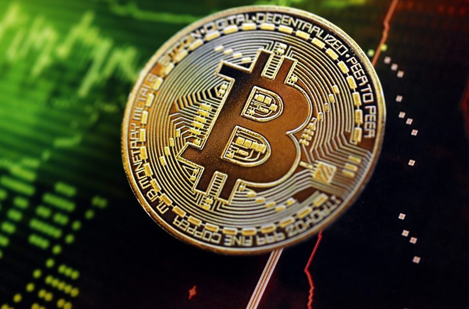 Eine physische Darstellung eines Bitcoins liegt auf einem digitalen Hintergrund mit grünen und roten Markttrendlinien und Kurscharts.
