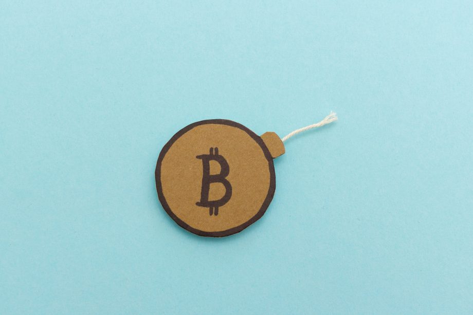Ein runder, brauner Papierausschnitt mit einem fetten Bitcoin-Symbol und einer Schnur, die wie eine Bitcoin-Bombe aussieht, ist auf einem hellblauen Hintergrund platziert.