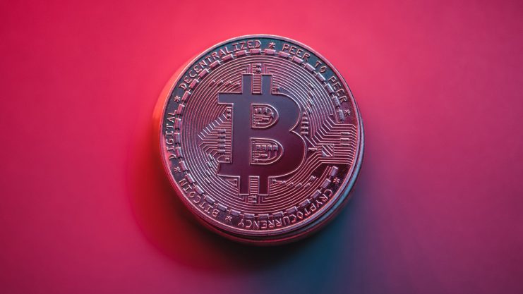 Eine physische Bitcoin-Münze ist auf einem Hintergrund mit rosa und lila Farbverlauf abgebildet und symbolisiert die Wirkung der digitalen Währung.