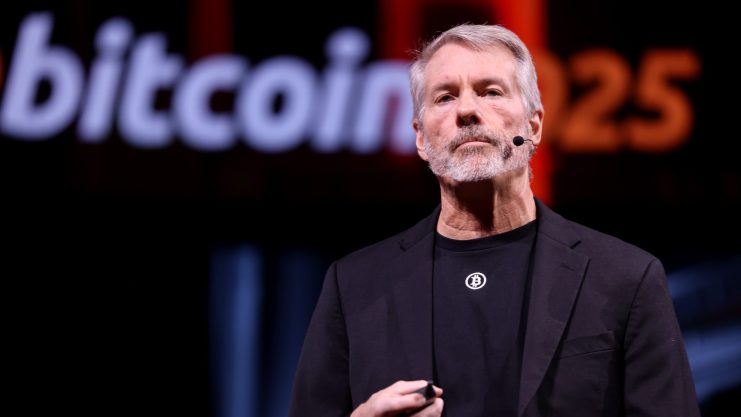 Ein Mann mit einem Headset-Mikrofon steht auf der Bühne vor einem verschwommenen "Bitcoin 2025"-Schild, trägt ein schwarzes Hemd und einen Blazer und ist bereit, über die Zukunft von Bitcoin zu sprechen.