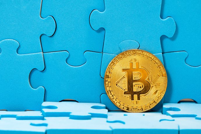 Eine goldene Bitcoin-Münze steht aufrecht vor einem Hintergrund aus blauen Puzzleteilen, von denen einige unvollständig oder unten abgetrennt sind und symbolisieren, wie Bitcoin in das komplexe Puzzle der modernen Finanzwelt passt.