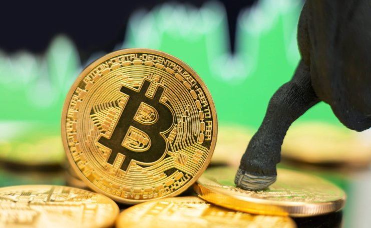 Eine goldene Bitcoin-Münze steht aufrecht auf einem Stapel ähnlicher Münzen, ein Stierhuf berührt den Bitcoin und im verschwommenen Hintergrund ist eine grüne Grafik zu sehen, was den kommenden Bullrun andeutet.