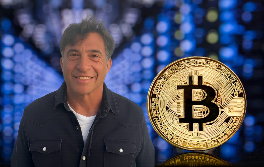 Ein Mann in einem dunklen Hemd steht lächelnd vor einem unscharfen digitalen Hintergrund, neben einer großen goldenen Bitcoin-Symbol-Münze.