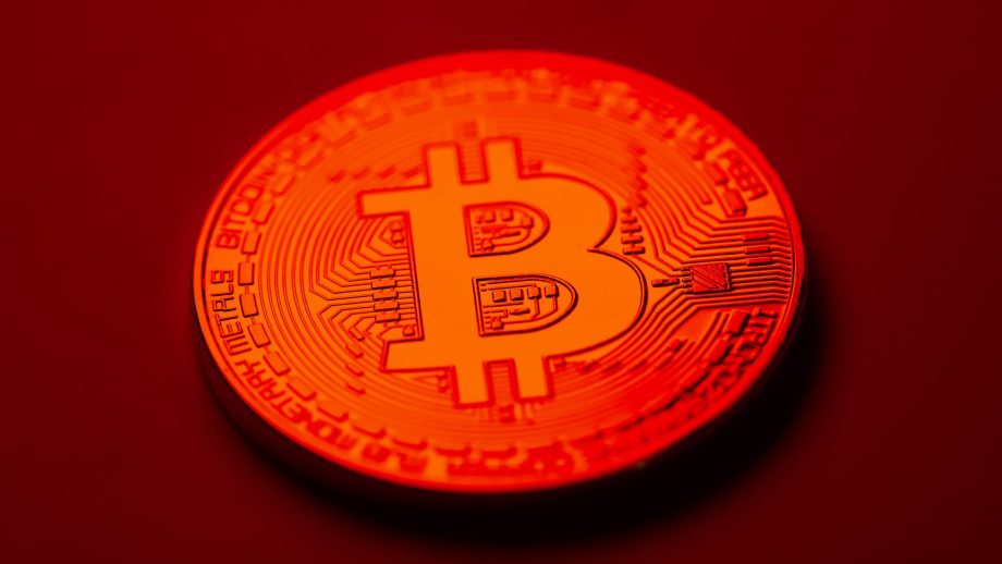 Eine Nahaufnahme einer physischen Bitcoin-Münze, die mit rotem Licht beleuchtet wird und das ikonische Bitcoin-Symbol sowie verschlungene kreislaufähnliche Muster zeigt.
