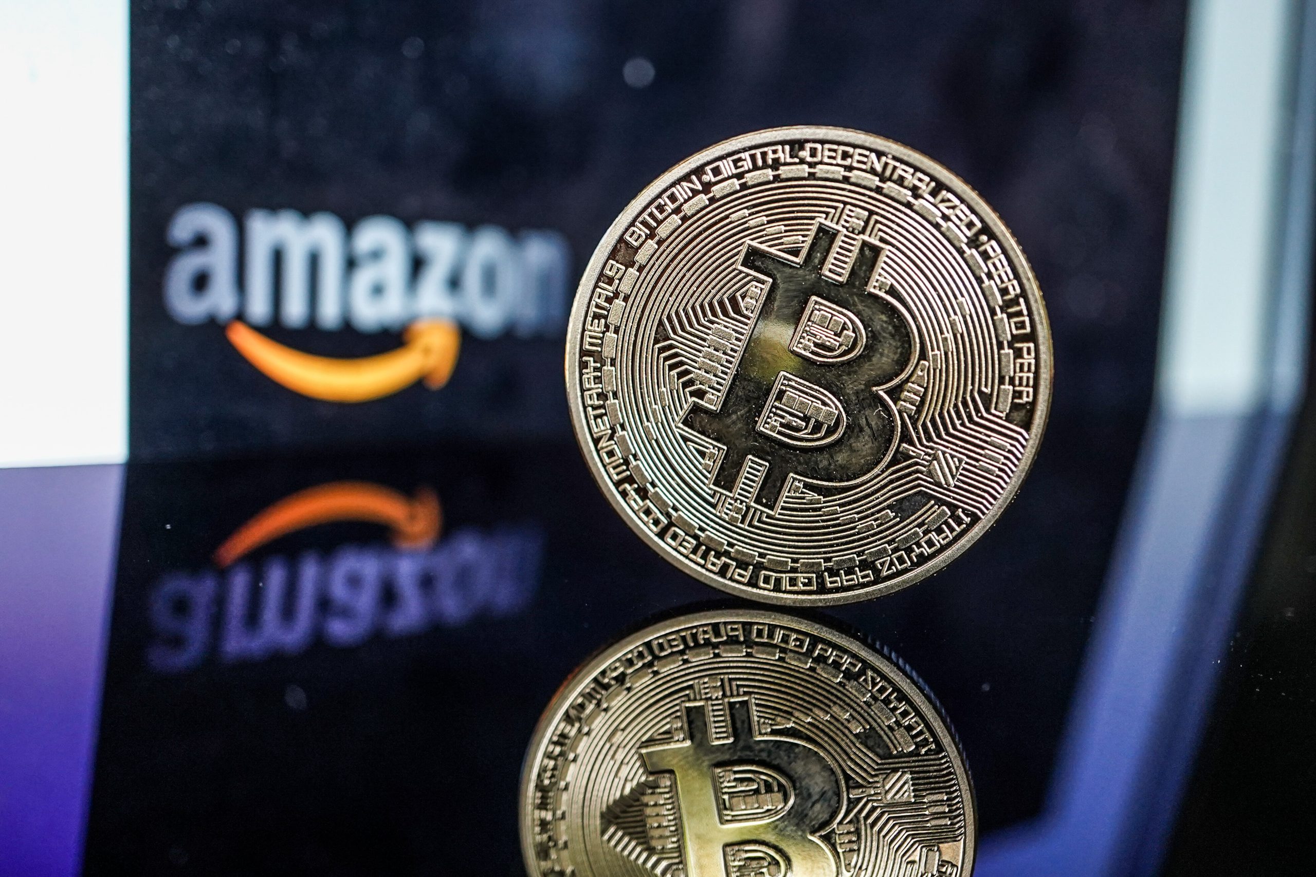 Eine physische Bitcoin-Münze steht aufrecht auf einer spiegelnden Oberfläche, mit dem verschwommenen Amazon-Logo im Hintergrund, um die wachsende Präsenz von Bitcoin im digitalen Handel hervorzuheben.
