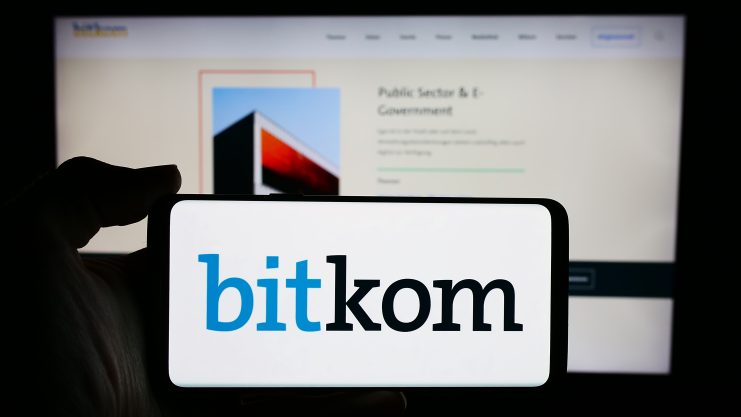 Eine Hand hält ein Telefon mit dem Bitkom-Logo vor einem verschwommenen Computerbildschirm, der eine Bitkom-Webseite zeigt.